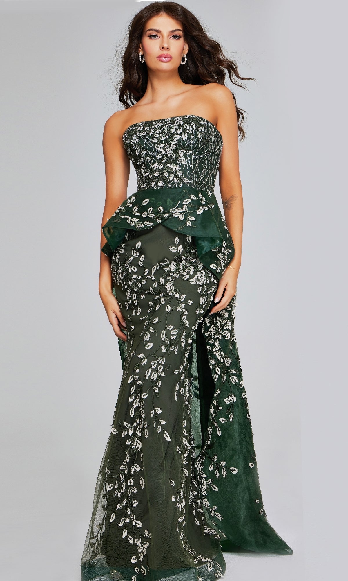 A woman wears the Jovani 37599, a long dark green formal dress with silver leaf embroidery and peplum detail, standing against a plain background.